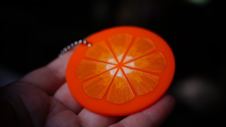 May include: A bright orange keychain shaped like a slice of orange. The orange slice has a white center and is detailed with lines to resemble the segments of an orange.