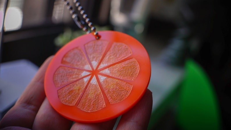 May include: Orange slice keychain made of bright orange plastic with a silver chain. The keychain is shaped like a slice of orange with a textured surface.