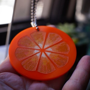 May include: A round orange pendant necklace with a detailed orange slice design. The pendant is made of acrylic and has a shiny finish.