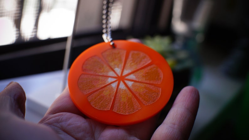 May include: A round orange pendant necklace with a detailed orange slice design. The pendant is made of acrylic and has a shiny finish.
