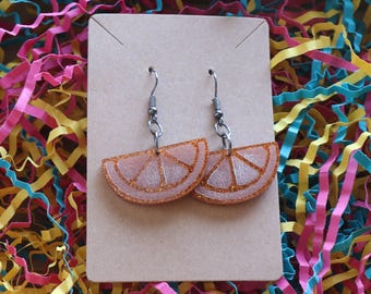 May include: A pair of dangle earrings shaped like orange slices, crafted from translucent orange resin with embedded glitter. The earrings feature silver-toned metal hooks and are attached to a rectangular brown cardboard display card.