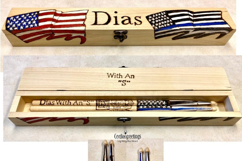 May include: A wooden box with a hinged lid, decorated with American and Thin Blue Line flags. The box contains two wooden drumsticks with the text "Dias With An "S"" engraved on them. The box is engraved with the text "Dias" and "With An "S"".