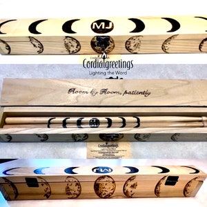 May include: A wooden box with a hinged lid, decorated with a moon phase design and the initials "MJ". The box contains two wooden drumsticks. The box is engraved with the text "Room by Room, patiently".
