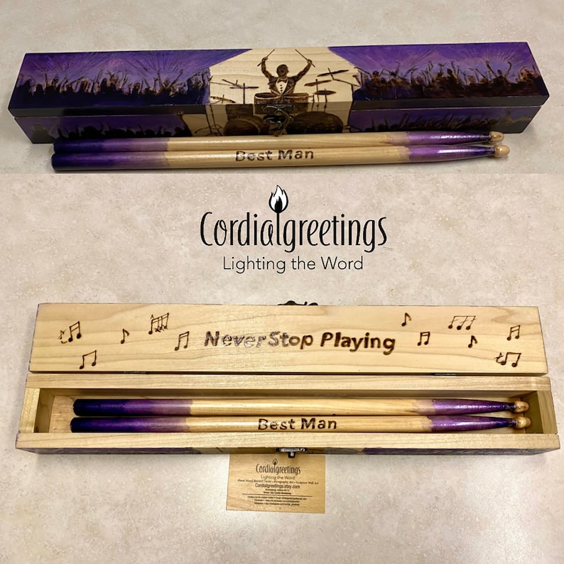 May include: A wooden box with a purple and black image of a drummer on the lid. The box contains two wooden drumsticks with purple tips. The box is engraved with the text "Never Stop Playing" and "Best Man".
