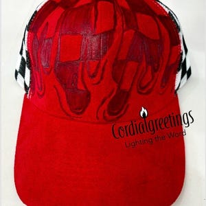 May include: A red baseball cap featuring a hand-painted flame design in darker red tones on the front panels. The sides of the cap have a black and white checkered pattern. The text Cordialgreetings Lighting the Word is printed in black.