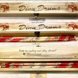 May include: A wooden box with a hinged lid and a latch. The box is engraved with the words "Dina Drums" and a drawing of a drum set. The box contains a pair of drumsticks with a musical note design.