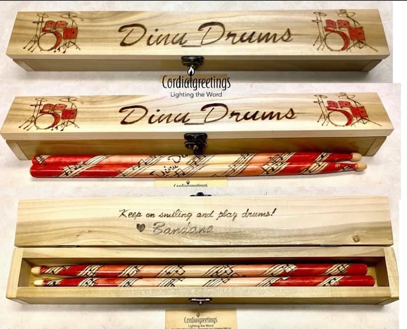 May include: A wooden box with a hinged lid and a latch. The box is engraved with the words "Dina Drums" and a drawing of a drum set. The box contains a pair of drumsticks with a musical note design.