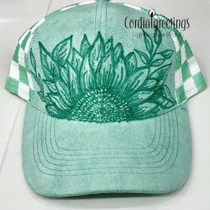 May include: A mint green baseball cap featuring a dark green line art illustration of a sunflower and foliage on the front panel. The cap has a white and mint green checkered mesh back and includes the text Cordialgreetings Lighting the Word.