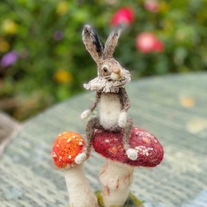 May include: A needle felted rabbit wearing a white collar sits atop a red and orange mushroom with white stems. The mushroom is sitting on a bed of green and brown felt.