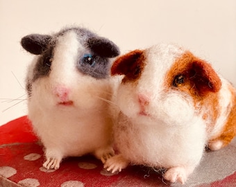 May include: Two handmade guinea pig figurines. One is gray and white, the other is brown and white. Both have pink noses and are sitting on a red polka dot surface. The figurines are made of soft, fluffy material.