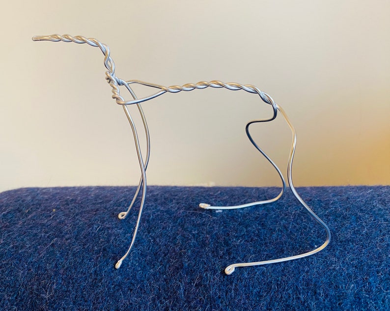 May include: A silver wire sculpture of a dog in a standing pose. The wire is twisted and bent to create the shape of the dog.