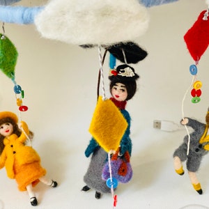 May include: A whimsical felt mobile featuring three figures suspended from a blue ring and a fluffy white cloud. Each figure holds a colorful kite. The figures are dressed in vintage-style clothing, with a yellow coat and skirt, a blue coat and gray skirt, and a gray suit.