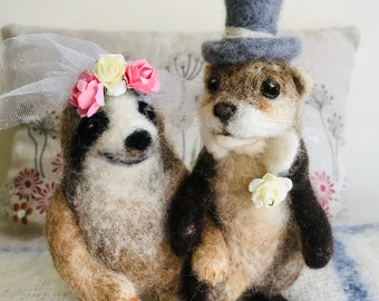 May include: Needle felted sloth and otter wedding cake toppers. The sloth wears a veil and floral crown, while the otter sports a top hat and boutonniere. The figures are tan, brown, and black, with delicate details. A unique wedding decoration.