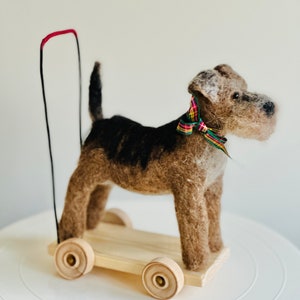 May include: A handmade felt dog toy with a wooden base and wheels. The dog has brown and black fur, a colorful plaid bow, and a red handle. The toy is designed to be pulled or pushed.