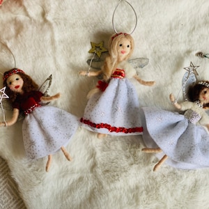 May include: Three handmade angel decorations. Each angel has a white dress with silver star details, silver wings, and a star-tipped wand. The angels have different hair colours and red accents on their dresses. Ideal for festive home decor.