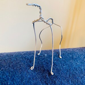 May include: A silver wire sculpture of a stylized animal with a long neck and four legs. The sculpture is standing on a blue surface.