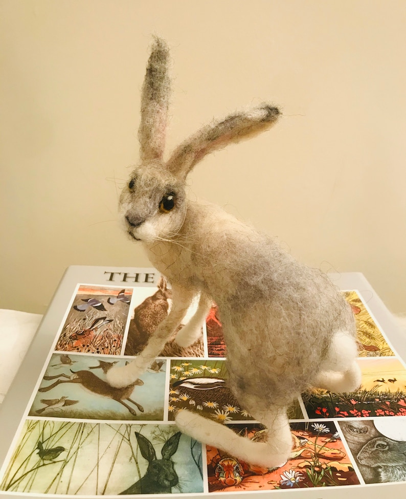 May include: A needle felted gray and white rabbit sculpture with brown eyes. The rabbit is sitting on a book with illustrations of animals and birds.