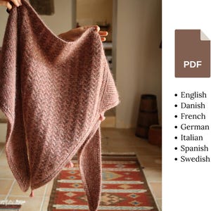 Peut inclure: Un châle triangulaire tricoté de couleur rose poudré présentant un motif à chevrons texturé sur tout le tissu. Le châle possède des extrémités pointues et de petits pompons aux coins. Une icône PDF marron affiche une liste de langues.