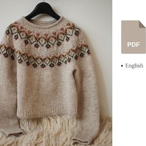 Pode incluir: Um suéter bege de malha com um jugo estampado em tons de marrom, vermelho e verde pendurado em um cabide preto. O suéter é exibido em um fundo branco e uma superfície fofa esbranquiçada. O ícone do arquivo PDF é visível.