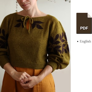 Può includere: Un maglione di lana verde con un motivo a foglie marroni sulle maniche e sul collo. Il maglione ha un laccio al collo.