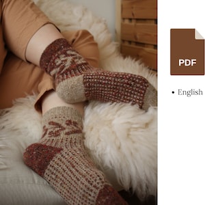 Può includere: Un paio di calzini di lana con un motivo marrone e rosso. I calzini sono indossati da una persona seduta su una superficie bianca e soffice.