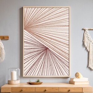 Puede incluir: Una impresión de arte abstracto enmarcada con un diseño geométrico de líneas marrones y rosadas que irradian desde un punto central. La impresión está colgada en una pared gris claro sobre una mesa de madera con una pared de macramé blanca a la derecha.
