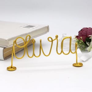 May include: A gold wire sculpture spelling out the word "aura" in cursive script. The sculpture is on a white surface, with a stack of books and a small floral arrangement in the background. The sculpture is approximately 6 inches wide.