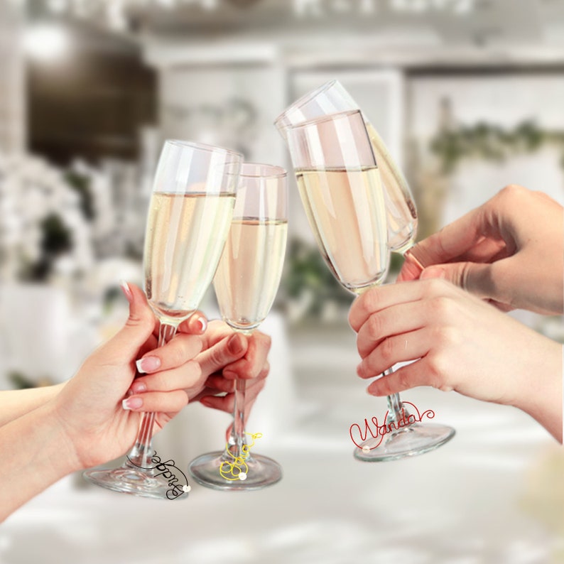 Op de afbeelding: Vier heldere glazen champagneflutes gevuld met mousserende wijn, omhoog gehouden in een toast. De flutes zijn versierd met draadnaamkaartjes met de tekst "Wanda", "Bride" en twee andere.