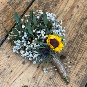 Puede incluir: Un pequeño ramo de flores secas con un solo girasol, aliento de bebé blanco y follaje verde. El ramo está atado con cordel y tiene dos alfileres con cabeza de perla.