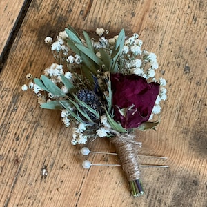 Puede incluir: Un pequeño ramo de flores secas con una sola rosa roja oscura, aliento de bebé blanco y follaje verde. El ramo está atado con cordel y tiene dos alfileres con cabeza de perla.