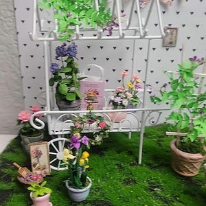 May include: Miniature white metal flower cart filled with colorful artificial flowers and greenery. The cart has a small pink bag with text, a small picture frame, and a decorative banner. The scene is set on a green, moss-like surface.