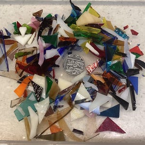 May include: A pile of multi-coloured glass shards, including red, blue, green, and white. A coin is included for scale. The glass pieces are of various shapes and sizes, likely for a craft project or mosaic art.