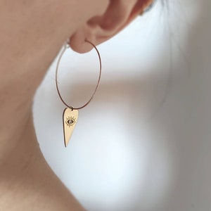 Puede incluir: Aretes de aro dorados con un dije colgante en forma de corazón con un diseño de ojo.