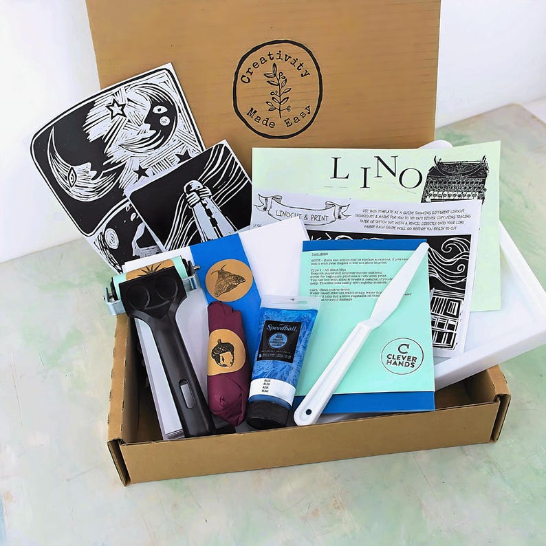 May include: A craft box filled with linocut and printmaking supplies. Includes a black and white print of a moon and stars, a Speedball blue ink tube, a white cutting tool, and other tools and materials. The box has the text "Creativity Made Easy".