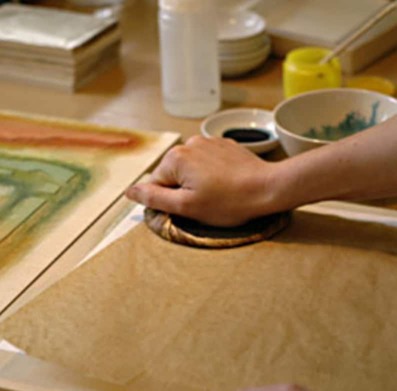 May include: A hand presses a brown wooden block onto a sheet of parchment paper. The paper is on a wooden table with a watercolour painting on it.