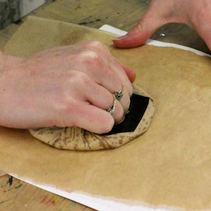 May include: A hand presses a brown and black patterned stamp onto a piece of parchment paper.