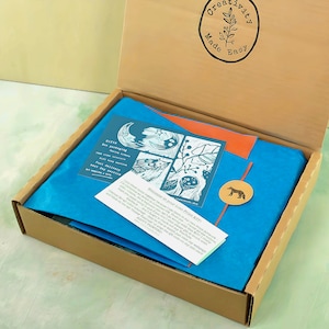 May include: A cardboard box filled with blue tissue paper, art prints, and a small round sticker with a fox illustration. The box also contains a card with the text "Welcome to your Lino Print Kit!" and the words "Creativity Made Easy" on the lid.