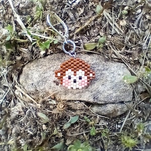 May include: A beaded monkey keychain with a silver-colored clasp. The monkey's face is made of small, colorful beads in shades of brown, white, pink, and black. The keychain is resting on a gray rock surrounded by grass and dirt.