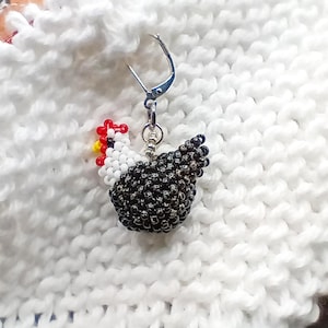 May include: A beaded chicken earring with a silver-tone leverback clasp. The chicken has a white head, red comb, yellow wattle, and a black beaded body. The earring is set against a white, textured background.