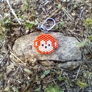 May include: A beaded keychain featuring a monkey design. The monkey's face is orange, white, and black, with a silver-colored metal keyring. The keychain is set against a natural background of a rock and foliage.