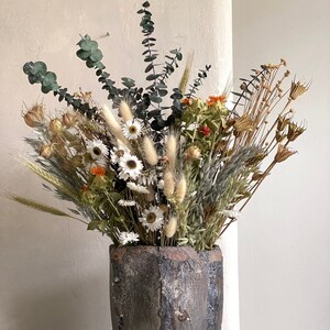 May include: A decorative vase made of weathered ceramic, holding a vibrant arrangement of dried flowers. The display features white daisies, green eucalyptus, and assorted dried grasses and seed heads. The vase has a dark, textured finish, enhancing the natural look.