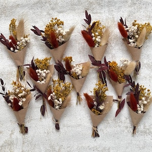 May include: A collection of twelve small bouquets wrapped in brown paper cones. Each bouquet features a mix of dried flowers including yellow yarrow, white baby's breath, dark red leaves, and fluffy tan bunny tail grass stems arranged together.