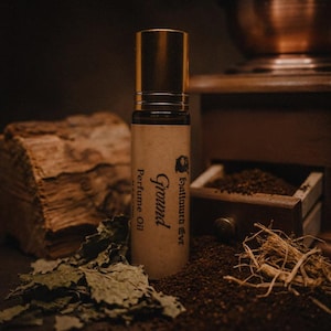May include: A cylindrical perfume oil roller bottle with a gold cap and a tan label featuring the text Hallowed Eve Ground Perfume Oil. The bottle is dark in color and stands upright, displaying clear black printed text on its paper label.