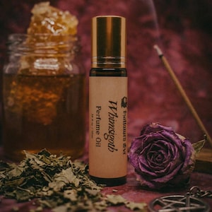 May include: A dark glass roller bottle of Whimsigoth Perfume Oil with a gold cap and a tan label reading .34 fl oz / 10ml. The bottle is surrounded by a jar of honey with honeycomb, dried green leaves, a purple rose, and a silver pentacle charm.