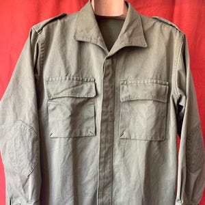 May include: Olive green military-style shirt with two chest pockets and buttoned cuffs. The long-sleeved shirt has shoulder epaulets and elbow patches. The shirt is hanging on a wooden hanger against a red background.