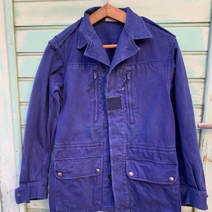 May include: A dark blue work jacket with multiple pockets and a zipper closure. The jacket has a button-up front and a patch on the left chest.