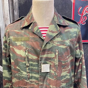 May include: A green, brown, and tan camouflage jacket with a zipper closure and two pockets on the front. The jacket has a patch on the left sleeve and a small pocket on the right chest.