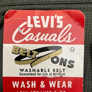 Può includere: Etichetta Levi's Casuals rossa e bianca con un grafico di fibbia di cintura dorata e il testo "WASHABLE BELT Guaranteed for Life of Garment WASH & WEAR LEVI STRAUSS & CO. SAN FRANCISCO, CALIF., U.S.A."