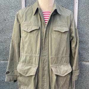 以下が含まれることがあります： オリーブグリーンのジャケット。前面に4つのポケットと襟が付いています。マネキンに着せられており、下に赤と白のストライプのシャツが見えます。ボタン留めとドローストリングウエストが付いています。