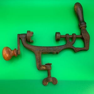 May include: An antique, rusty metal meat grinder with wooden handles and a clamp. The grinder has a crank on one side and a handle on the other. The clamp is used to secure the grinder to a table or counter.